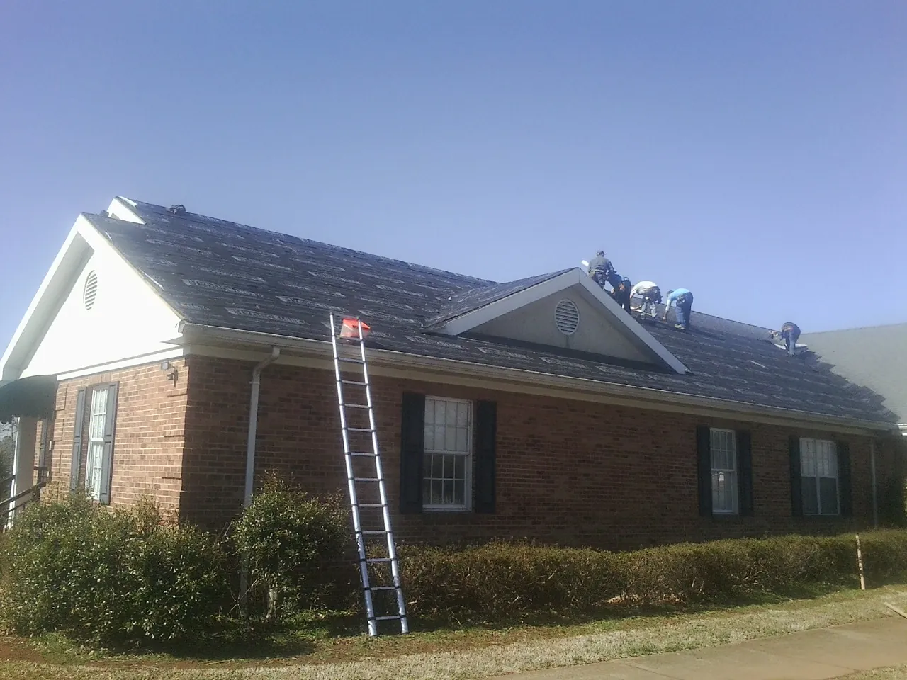 Crew tearing shingles off a brick ranch roof with a ladder set — Emergency Roof Repair work by our crew in Aynor
