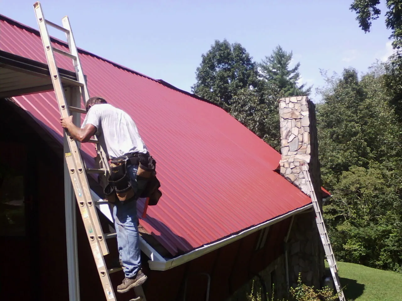 Roofer on a ladder beside a red metal roof and stone chimney — Soffit & Fascia Repair by Roofing Brothers in Aynor